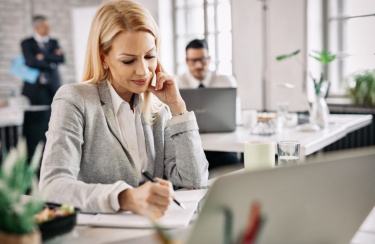 Eine Geschäftsfrau arbeitet an ihrem Schreibtisch und macht sich Notizen in ihrem Notizblock; im Hintergrund sind Personen zu sehen.