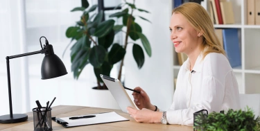 Lächelnde blonde junge Frau, die am Arbeitsplatz unter Verwendung der digitalen IPad sitzt